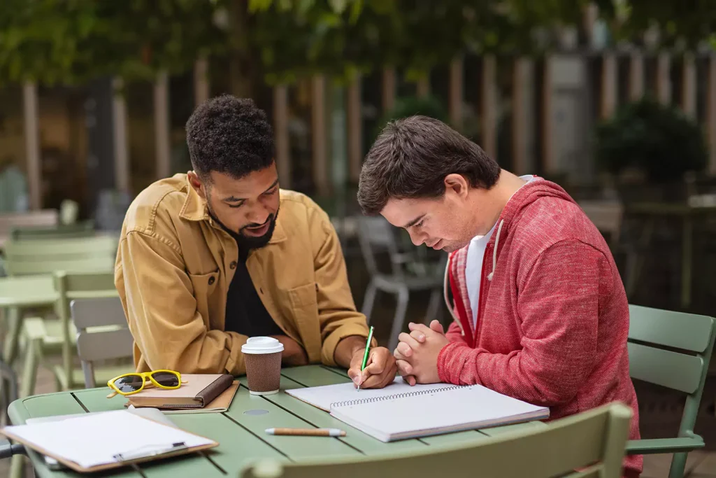 Disabled man with down syndrome writing in notepad with support worker